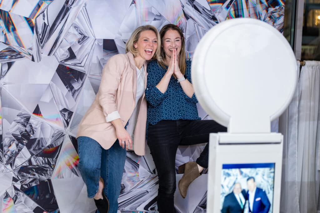 Two guests smiling and posing in front of a modern geometric backdrop using a white interactive photo booth at a corporate event.